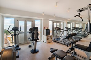 A gym with various exercise equipment, large windows, and a view of the city. - NH Collection Taormina (Taormina)