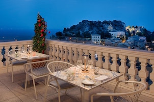 A terrace with a table set for dining, overlooking a coastal town and a mountain.