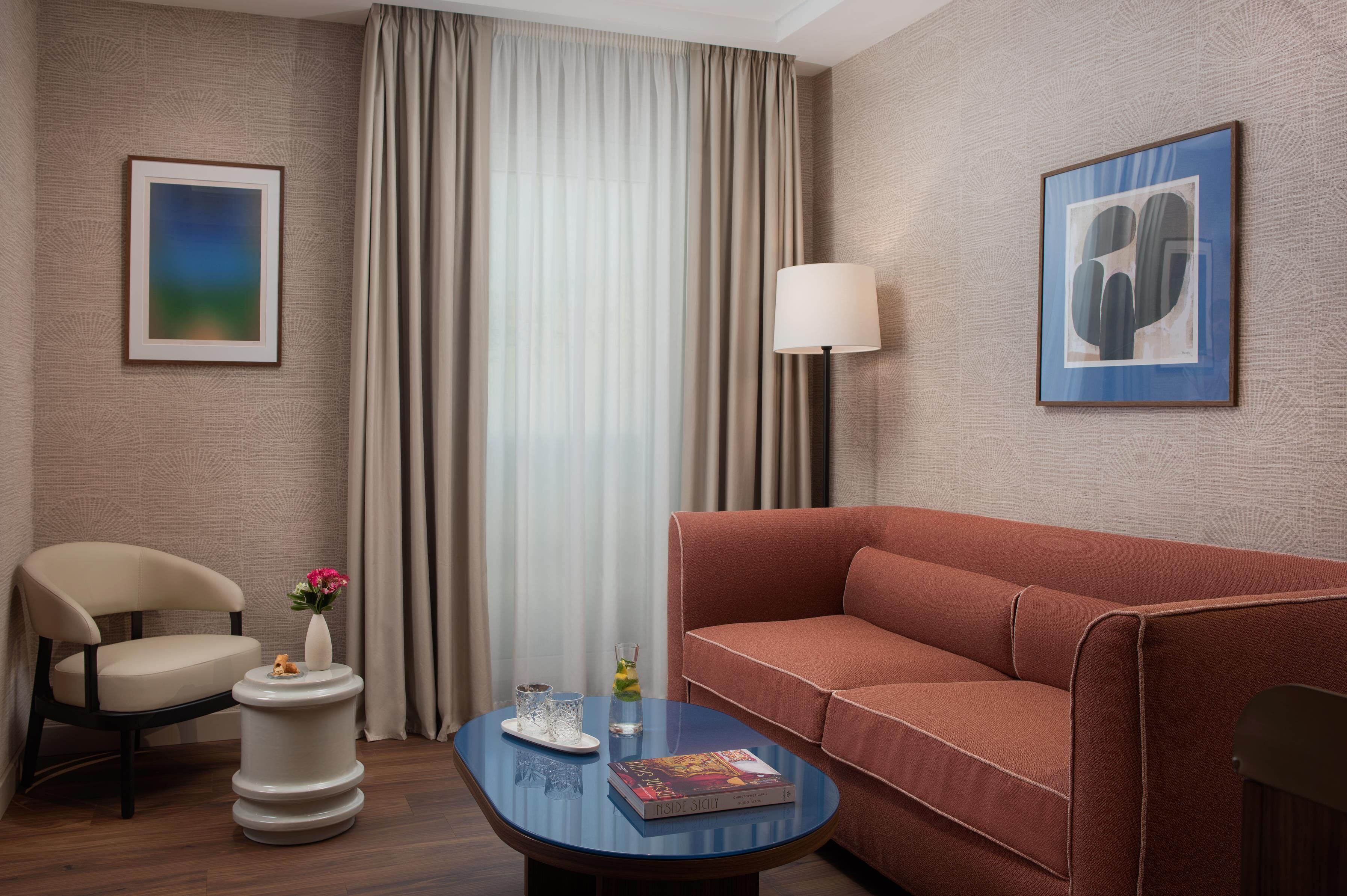 A hotel room with a sofa, a chair, a coffee table, a lamp, and two framed artworks on the walls.