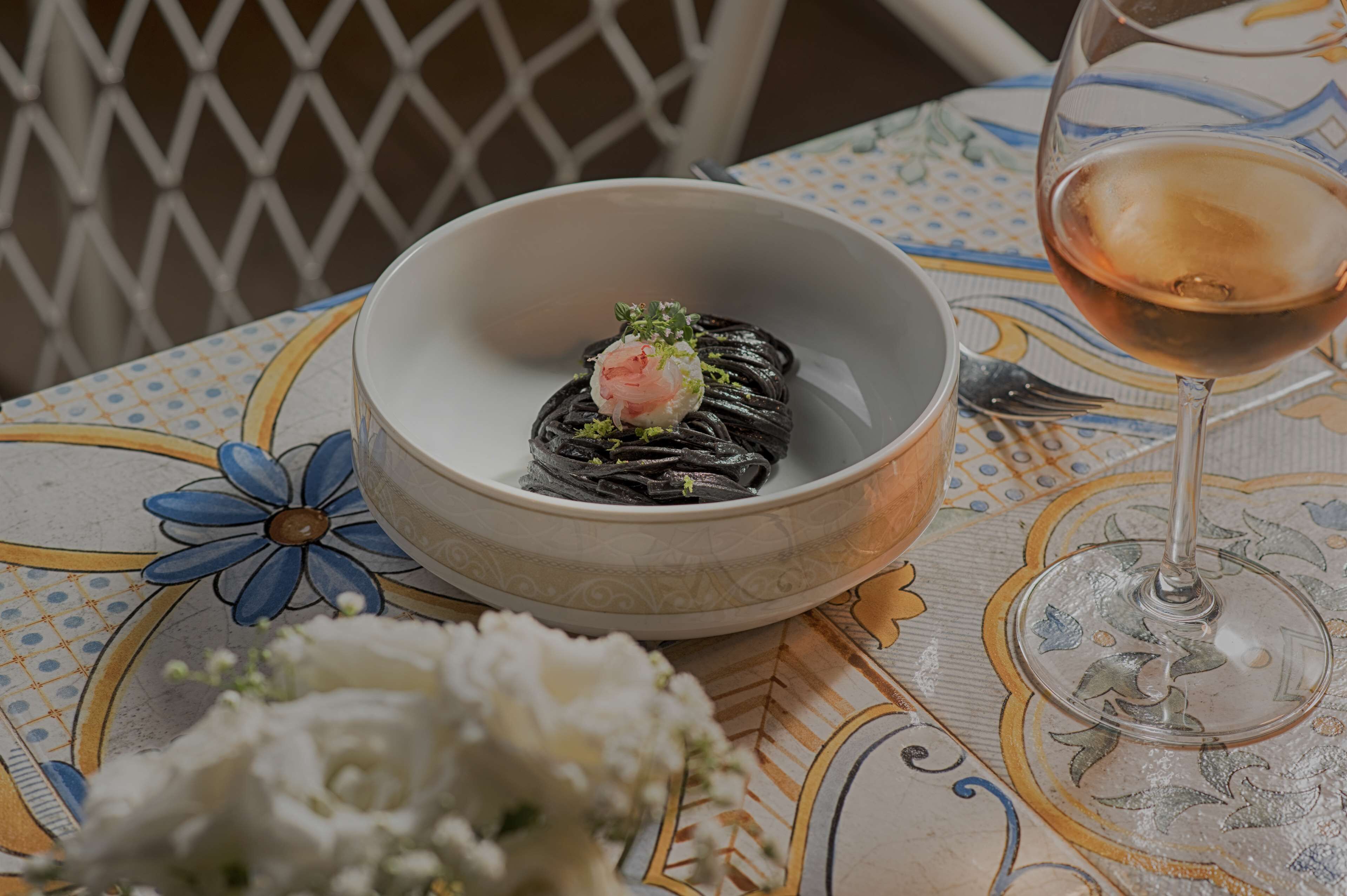 A table with a bowl of pasta, a glass of wine, and a floral arrangement.