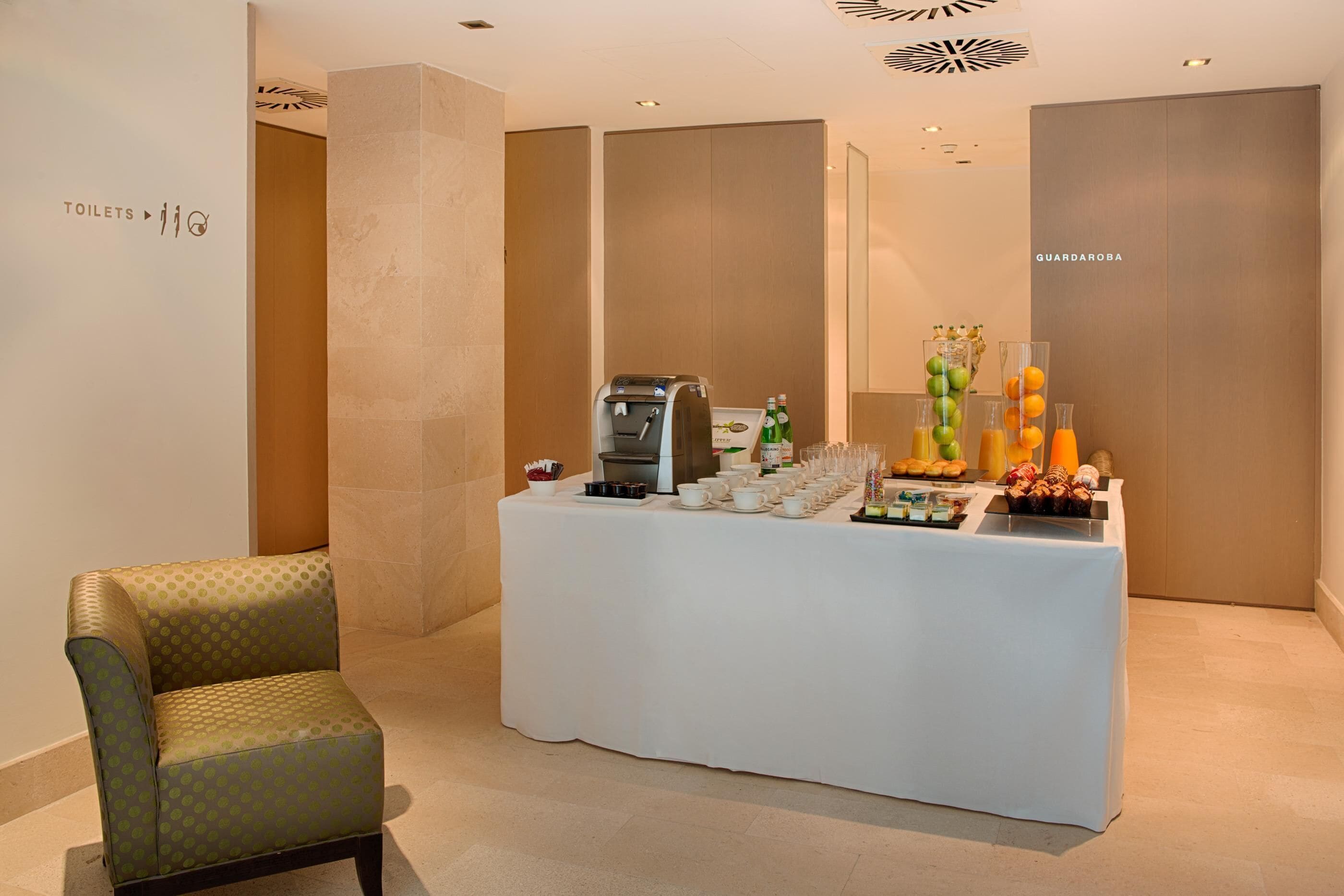 Quarto de hotel com balcão de café da manhã, máquina de café e diversas bebidas e doces.
