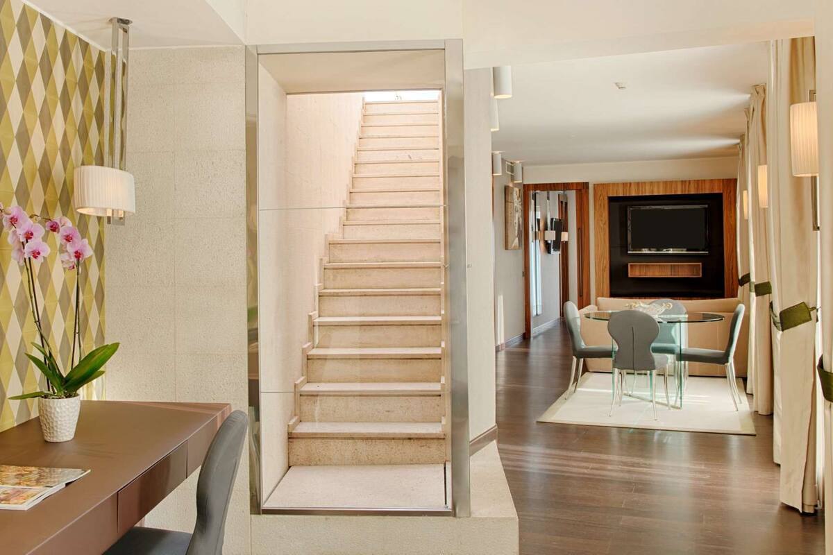 A modern interior with a staircase leading to a living area, a dining area with a table and chairs, and a TV mounted on the wall.