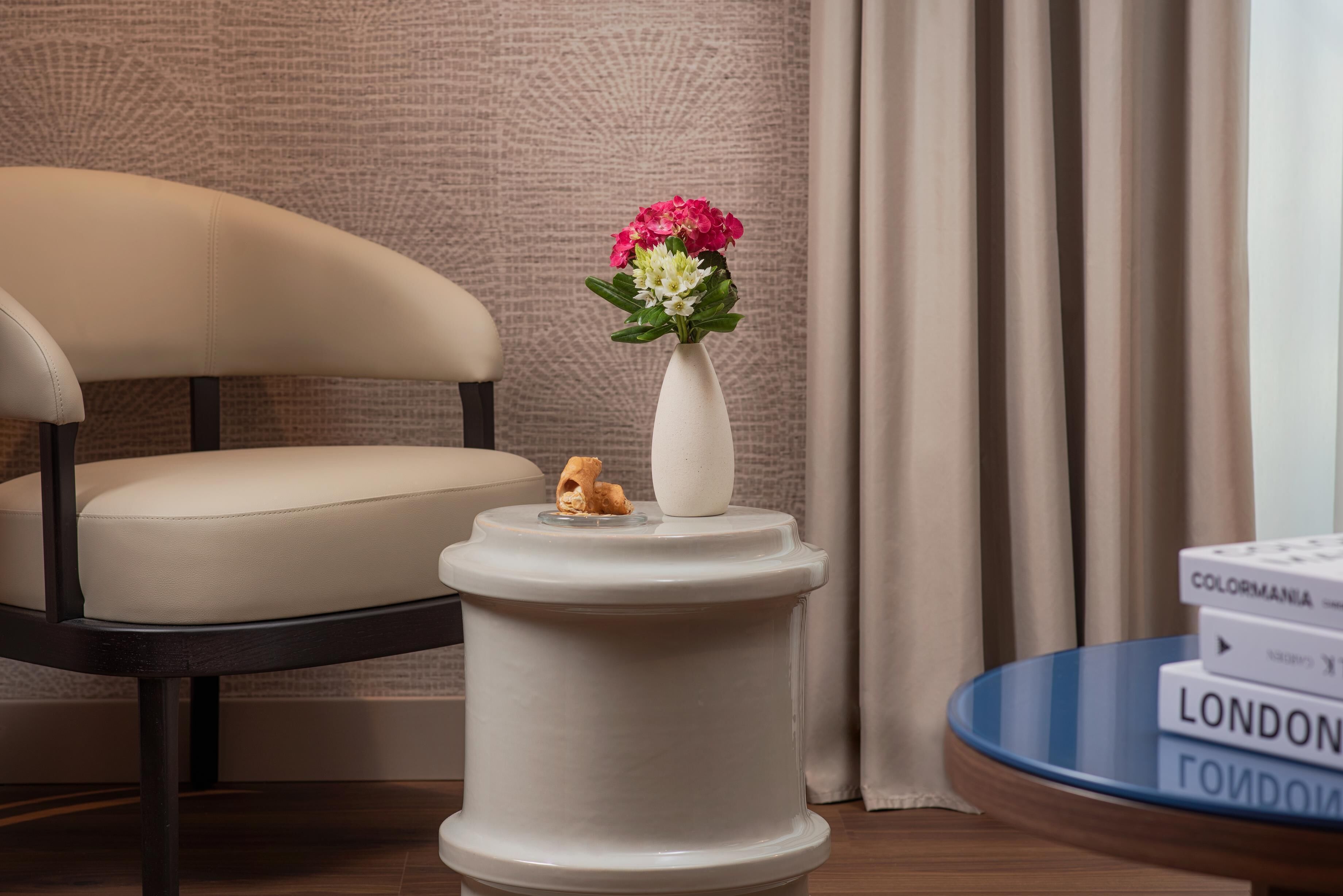 Una habitación con una silla, una mesita blanca con un florero y una pila de libros.