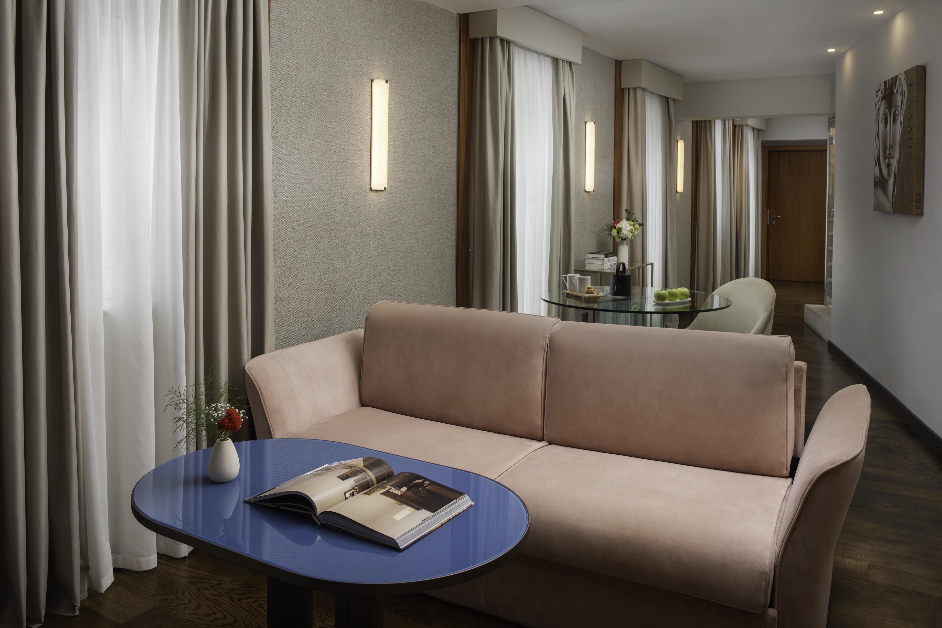 A modern hotel room with a beige sofa, a small round table with a magazine, and a glass coffee table.