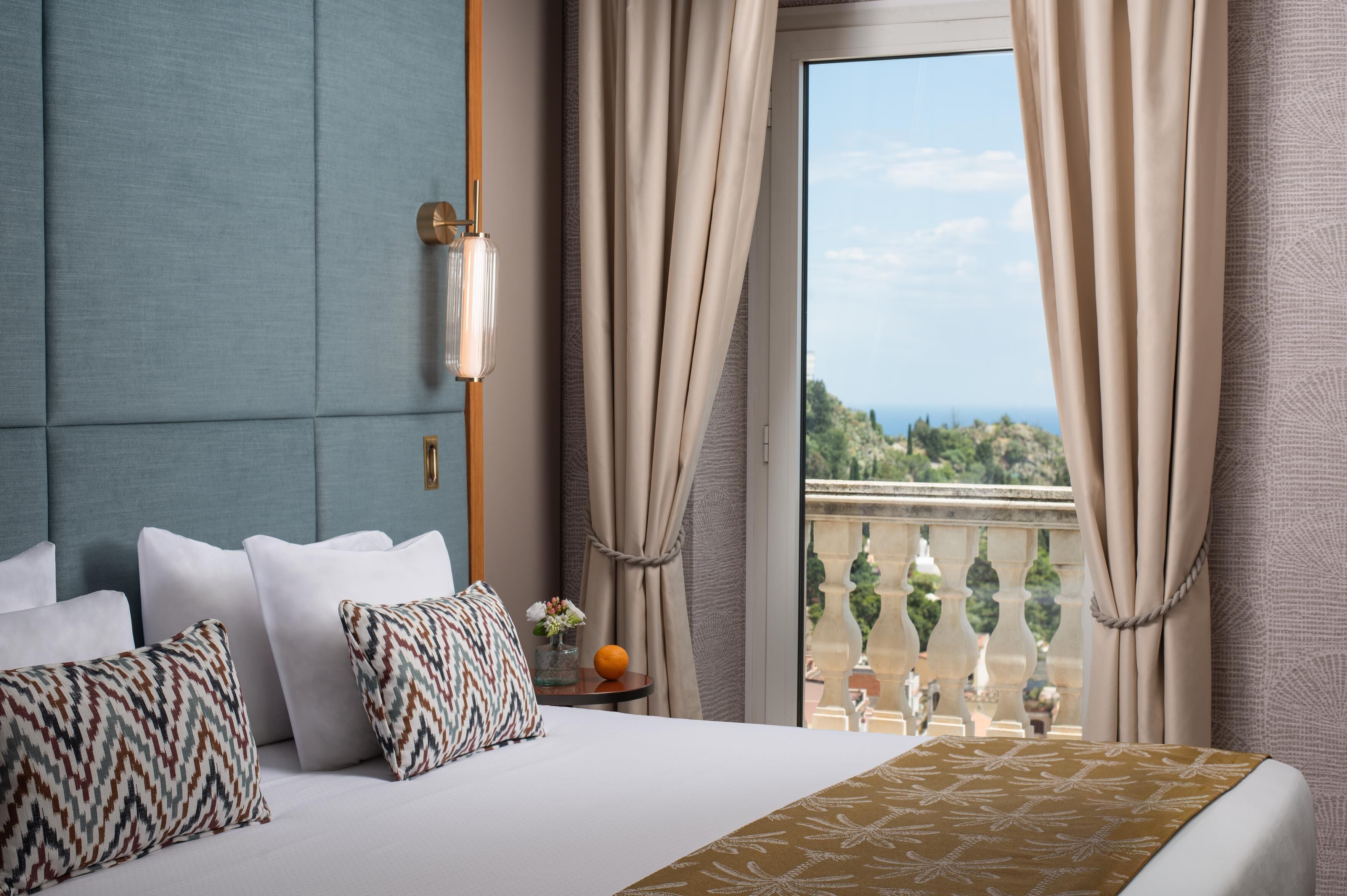 A hotel room with a bed, pillows, a nightstand, a vase with flowers, and a view of a balcony with a railing.