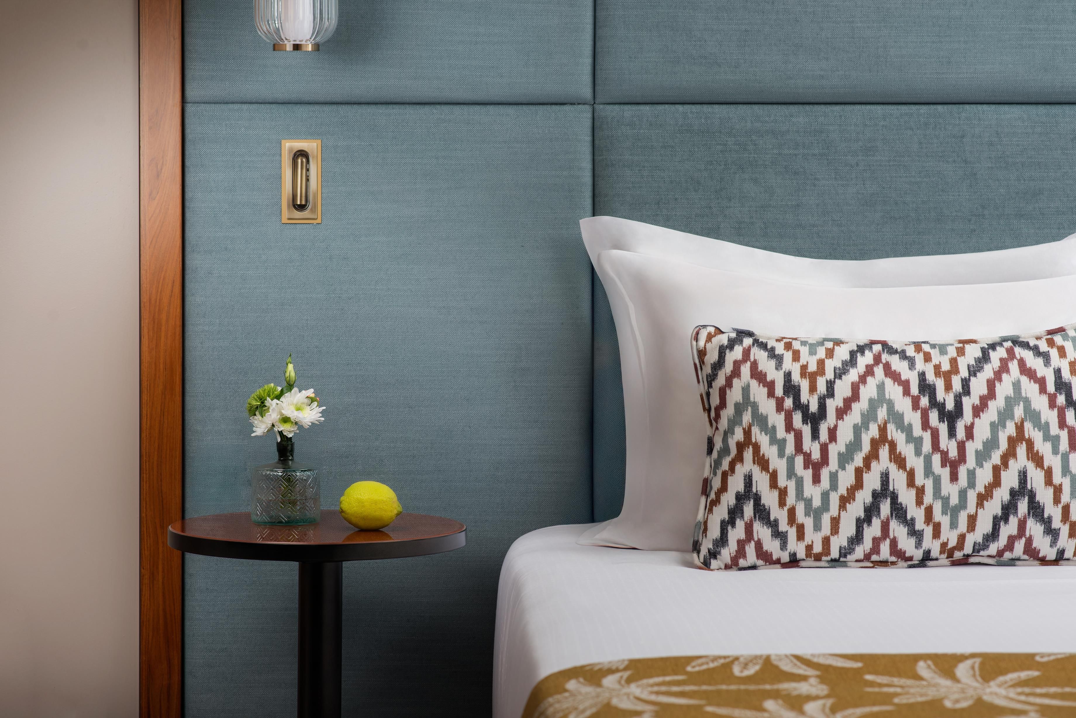 Hotelkamer met een bed, een klein tafeltje met een bloevas en een citroen, en een wandlamp.