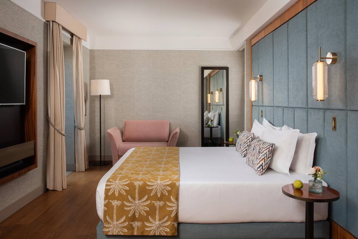 A hotel room with a bed, a chair, a TV, a mirror, and wall-mounted lights.