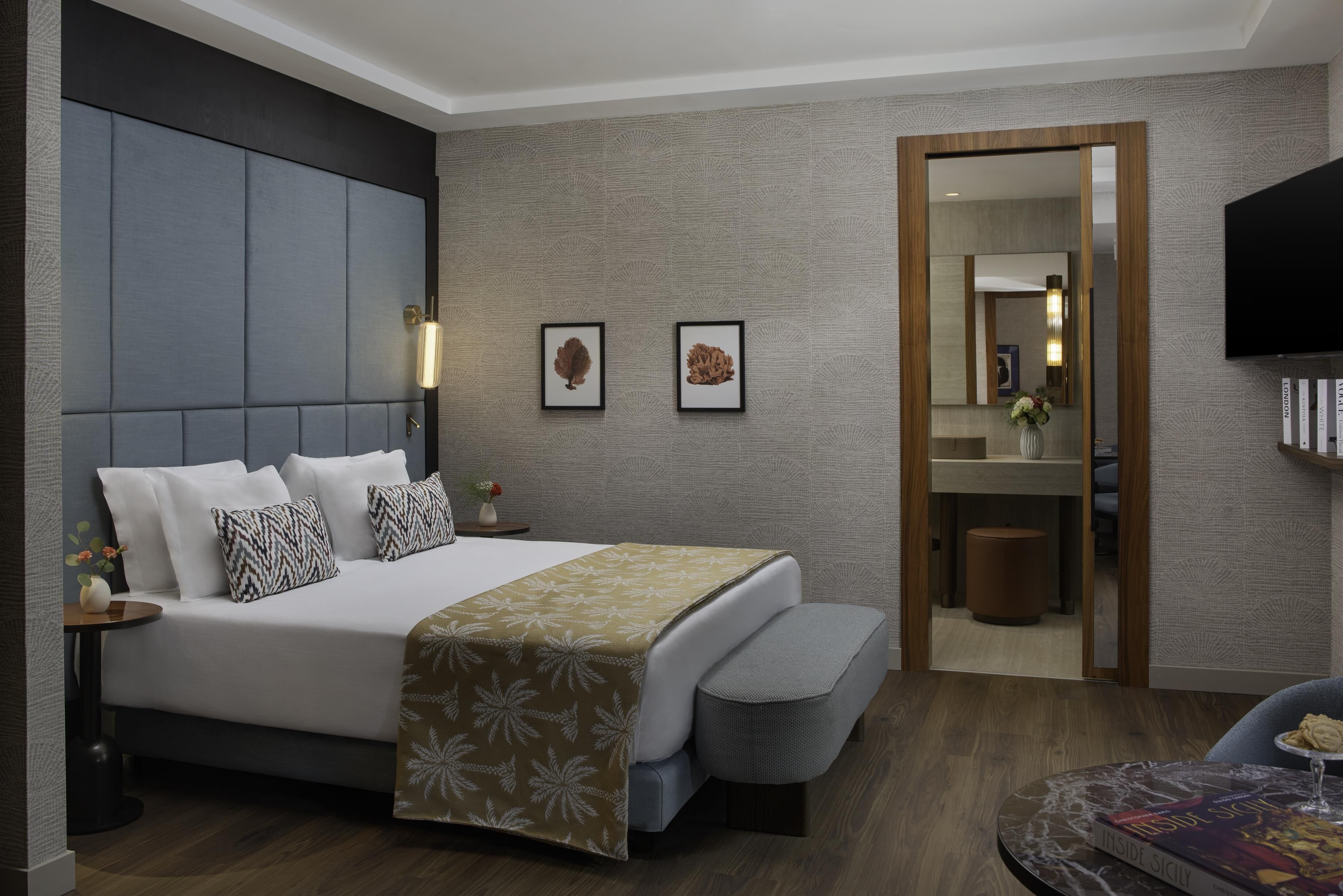 A modern hotel room with a large bed, bedside tables, a mirror, a television, and a small round table with a book and a glass.