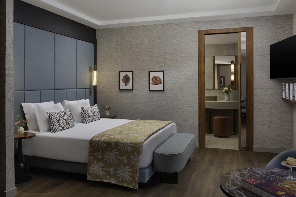 A modern hotel room with a large bed, bedside tables, a mirror, a television, and a small round table with a book and a glass.