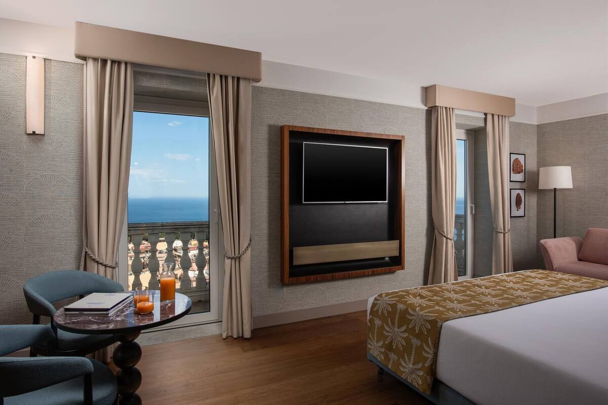 A hotel room with a bed, a desk with a chair, a TV mounted on the wall, and a view of the sea.