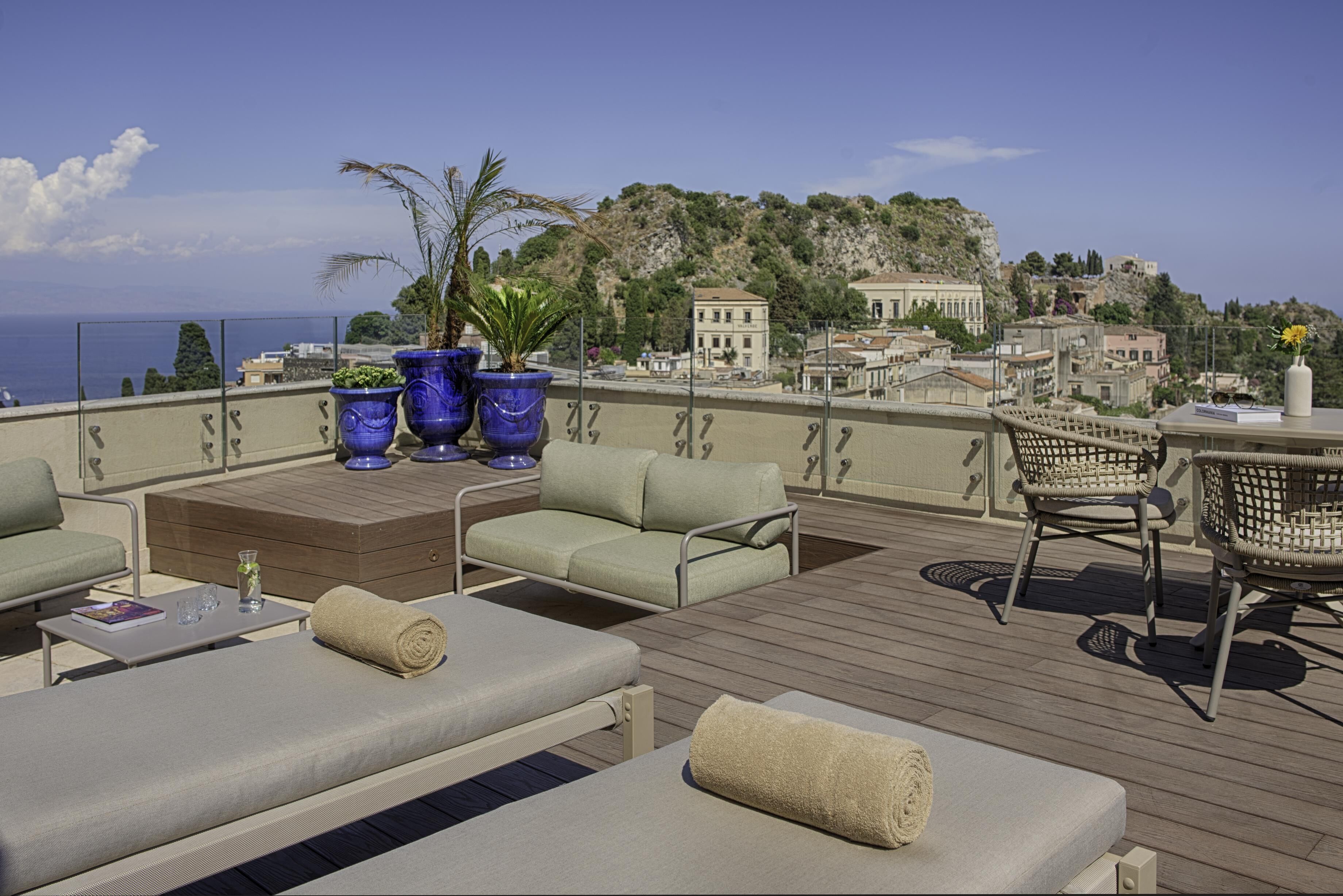 A rooftop terrace with outdoor furniture, potted plants, and a view of a coastal town.