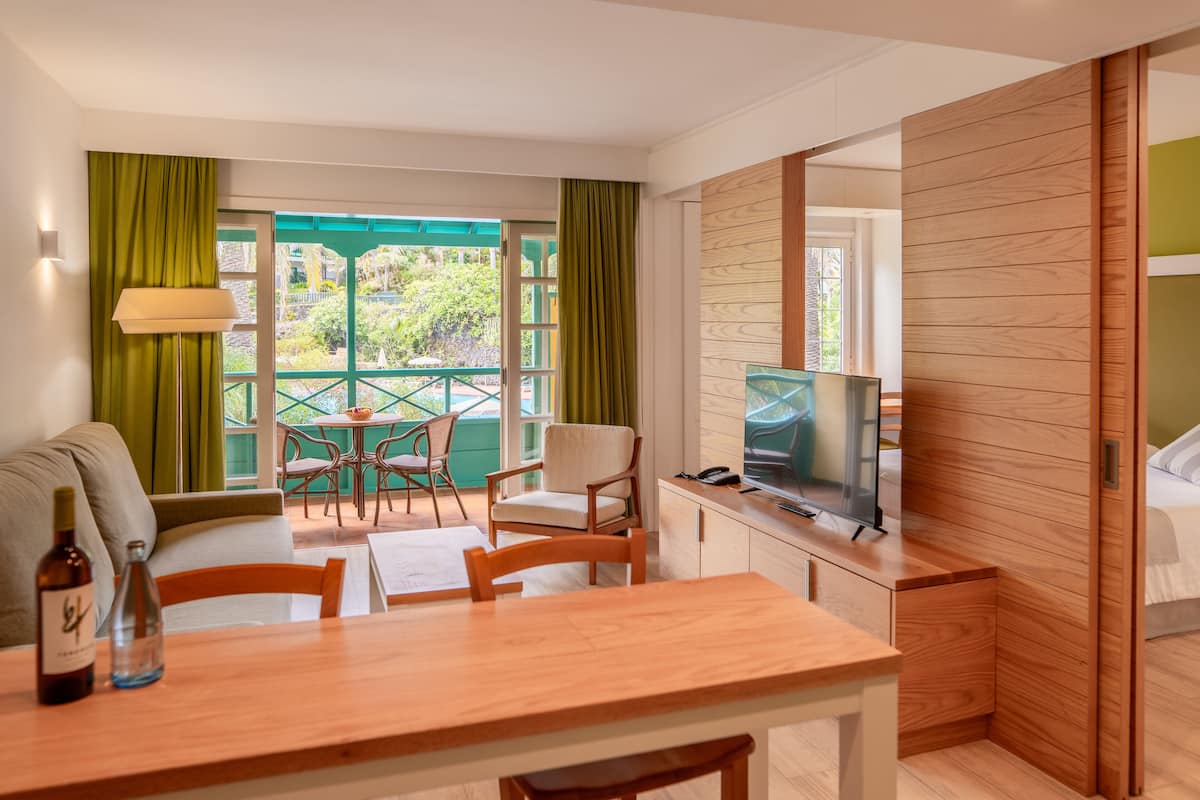 Ein modernes Zimmer mit einem Holztisch, Stühlen, einer Couch, einem Fernseher und einem Balkon mit Blick auf Grünflächen.