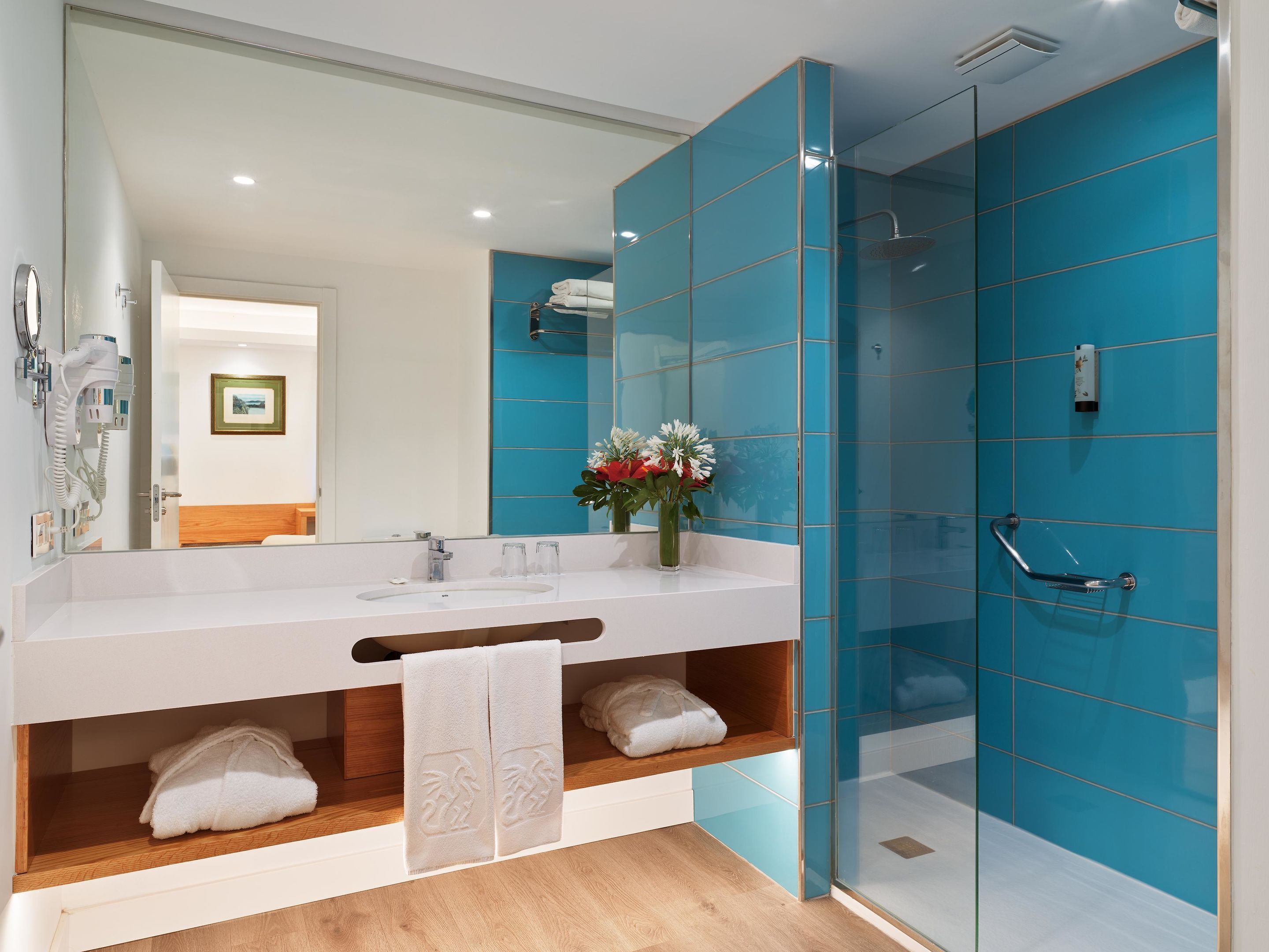 A modern bathroom with a large mirror, two sinks, a shower, and a wooden shelf with towels.