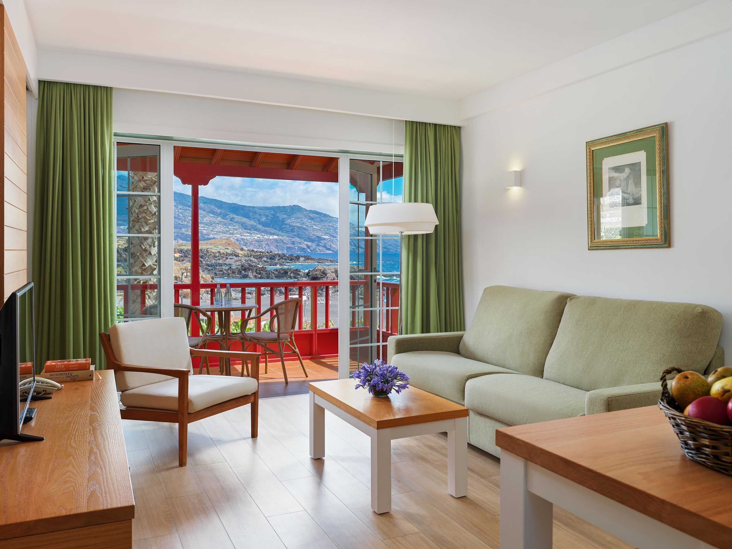 A living room with a sofa, a table, and chairs, a balcony with a view of the sea, and a framed picture on the wall.