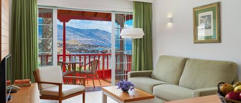A living room with a sofa, a table, and chairs, a balcony with a view of the sea, and a framed picture on the wall.