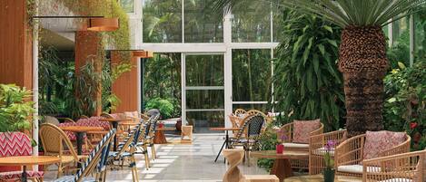 Un jardín interior espacioso con mobiliario de madera, amplias ventanas y abundante vegetación.