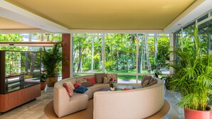 Amplia sala de estar con sofá curvo, una mesita de centro y mueble para televisión. Grandes ventanales ofrecen vistas a exuberante vegetación.