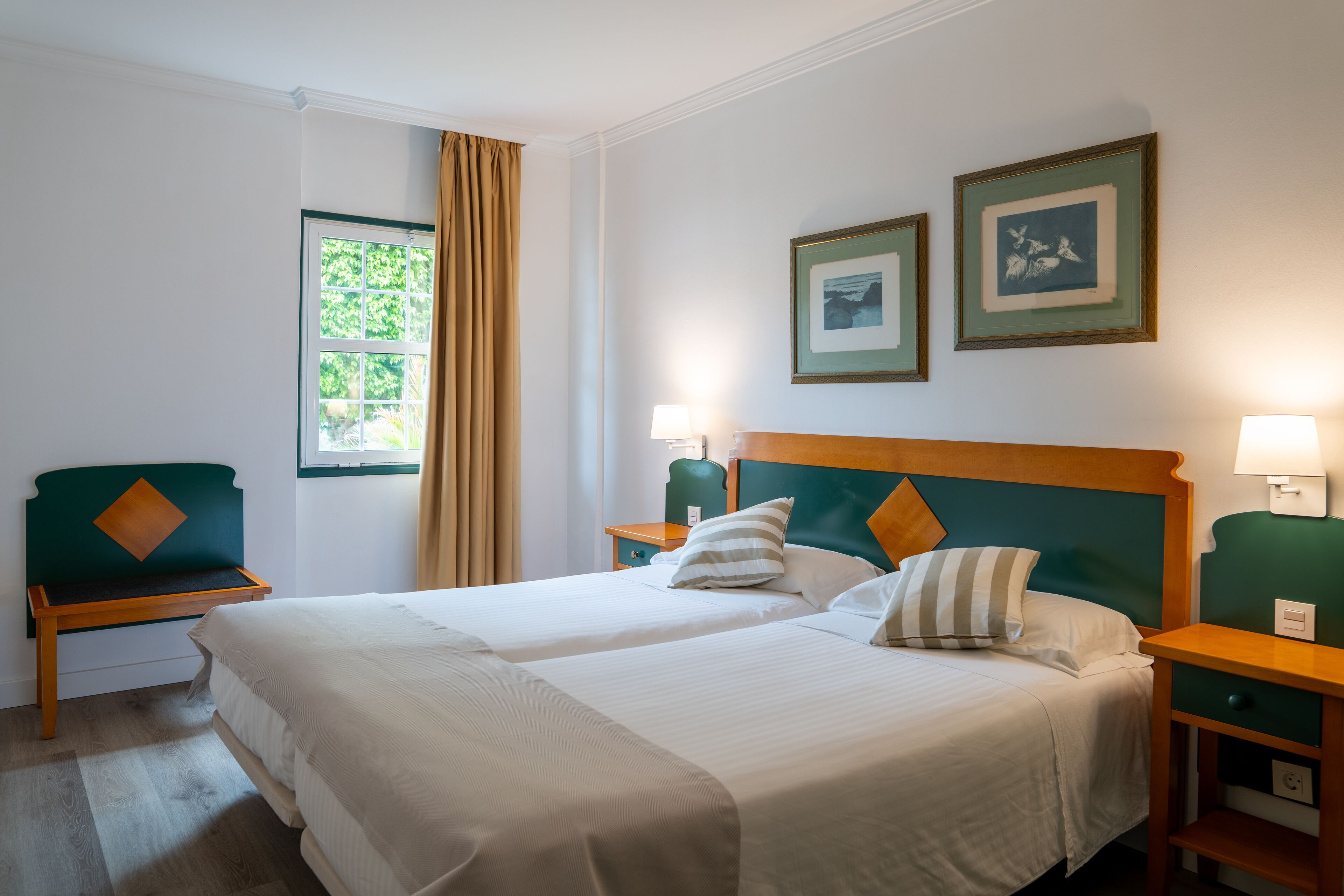 Une chambre d’hôtel avec deux lits, une table de chevet en bois, un fauteuil vert et une fenêtre avec des rideaux.