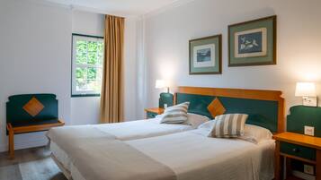 Une chambre d’hôtel avec deux lits, une table de chevet en bois, un fauteuil vert et une fenêtre avec des rideaux.