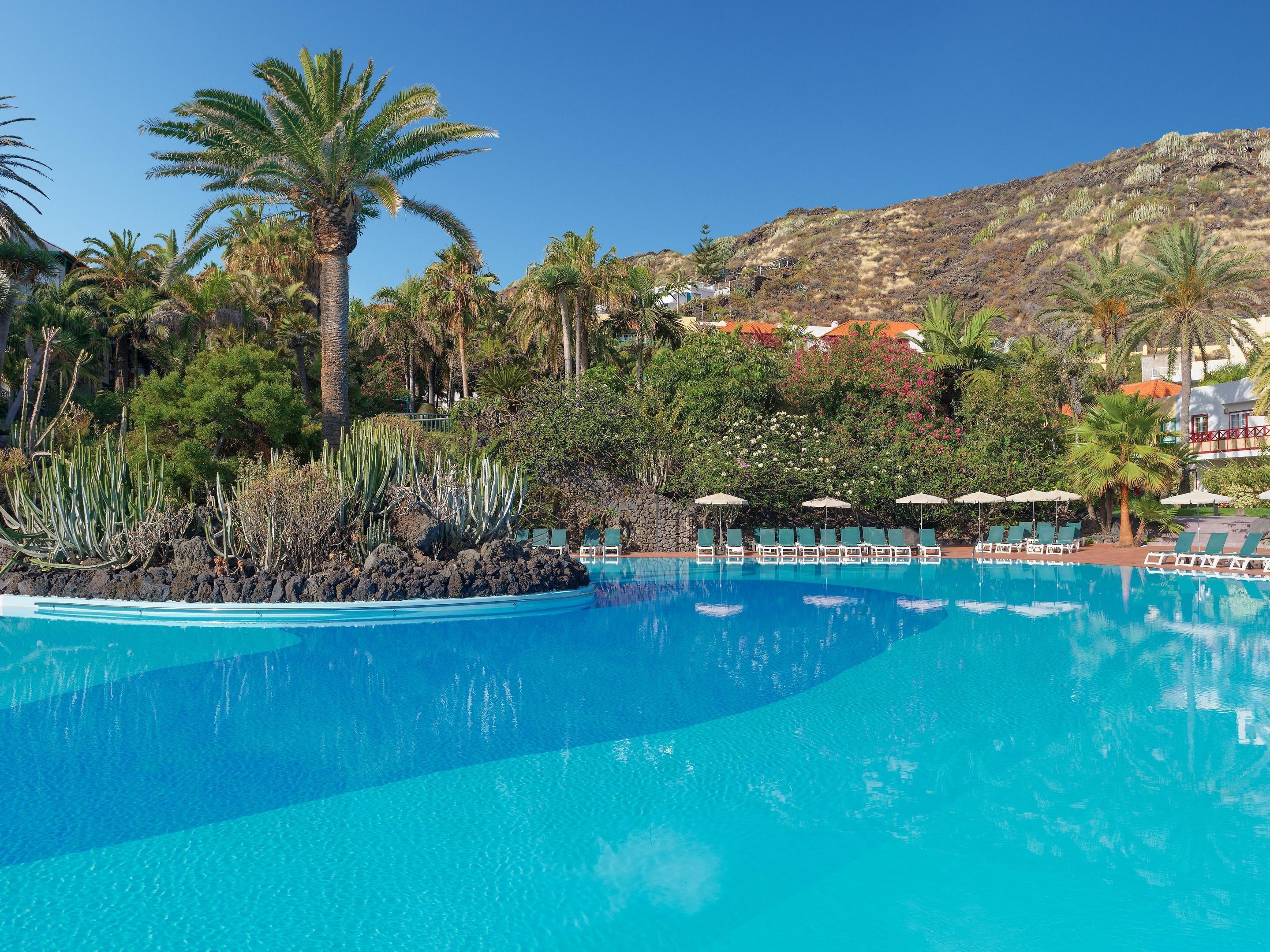 A clear blue pool surrounded by lounge chairs and palm trees, with a mountainous backdrop.