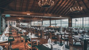Un restaurante espacioso con techos altos, vigas de madera y ventanas grandes que ofrecen vistas a las montañas. Las mesas están dispuestas ordenadamente con manteles blancos y sillas verdes.