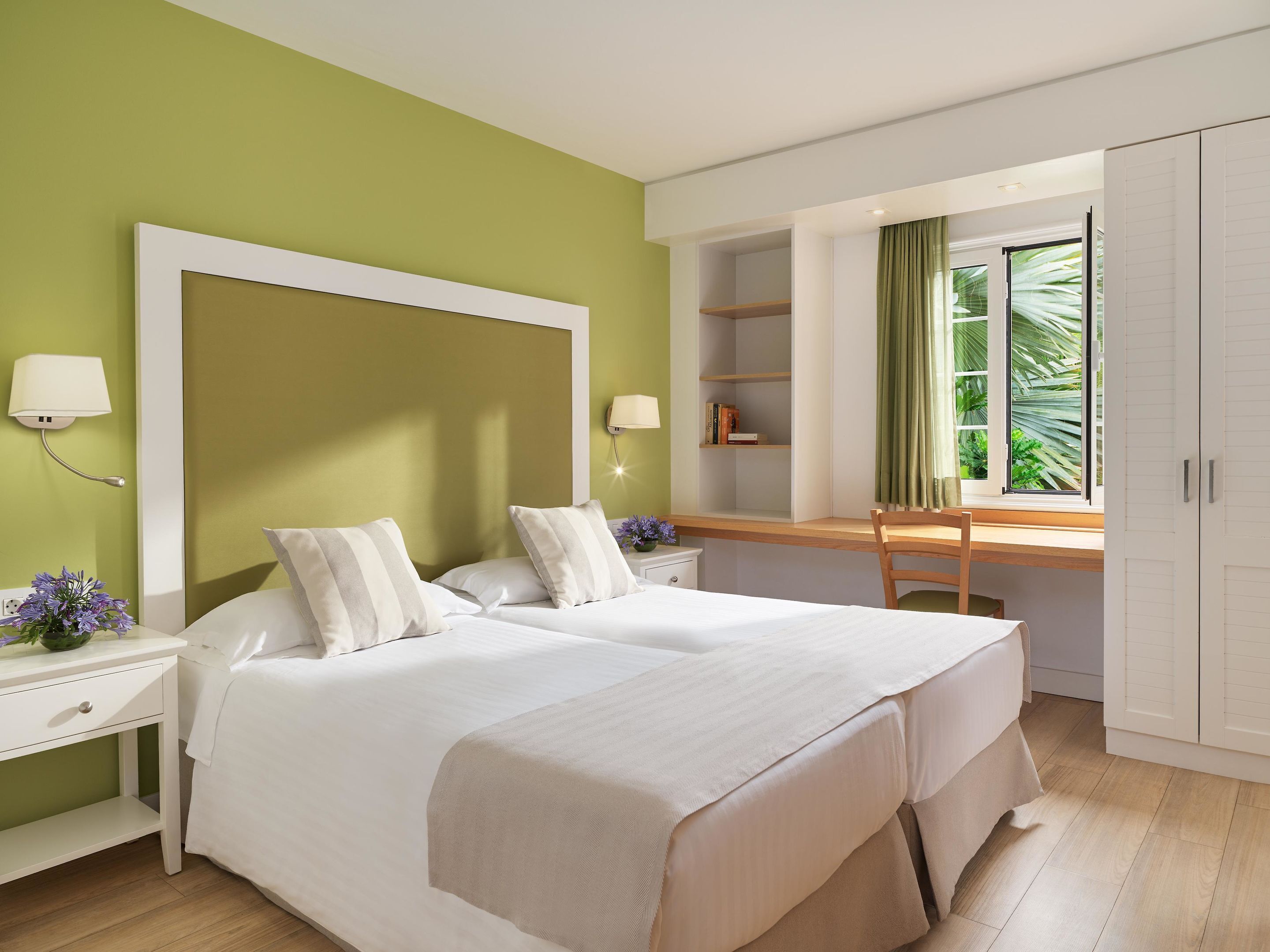 A hotel room with two beds, a desk, a chair, a wardrobe, and a window with curtains.