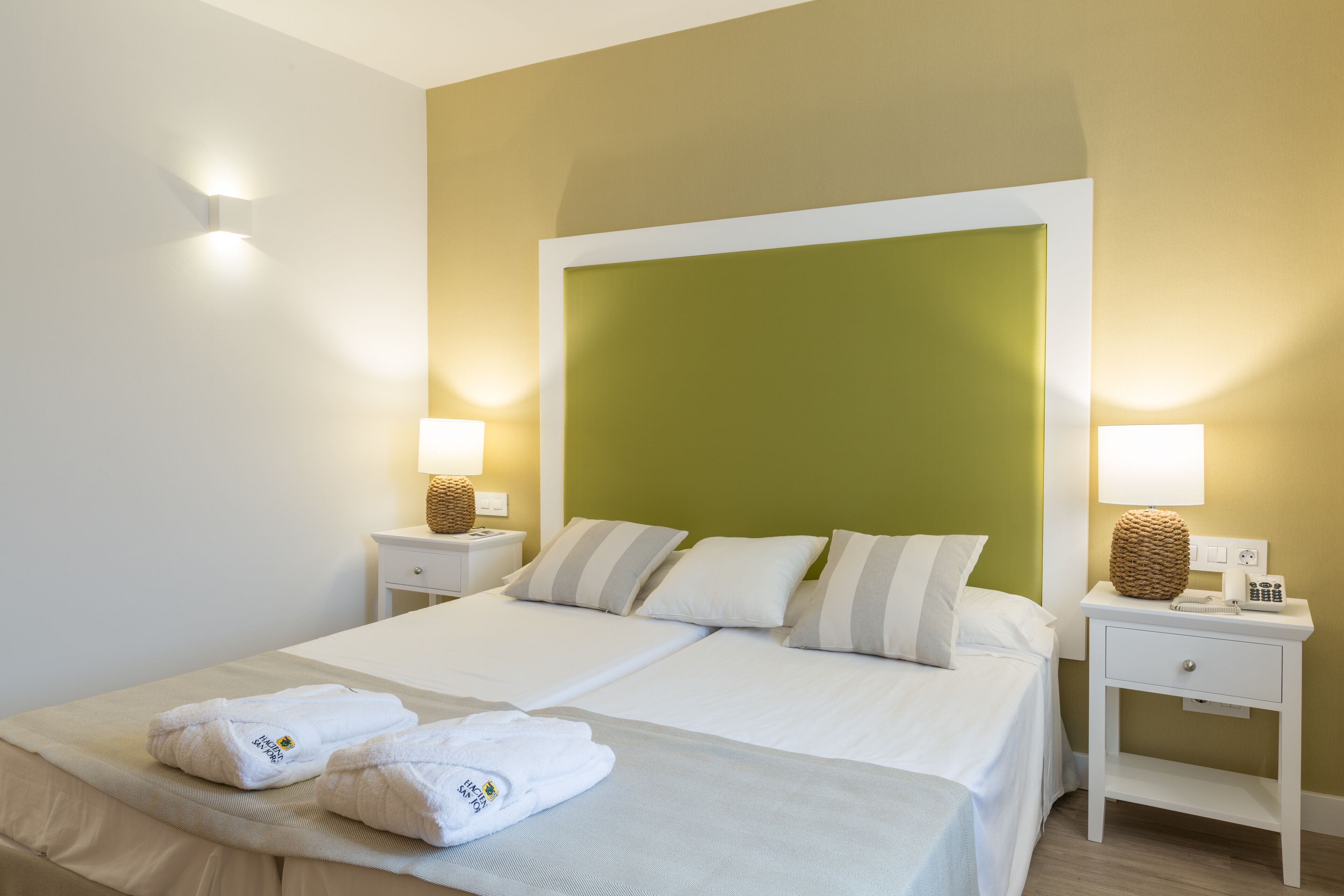 Cama doble con sábanas blancas y dos toallas, una mesita de noche con lámpara y una pared de acento verde.
