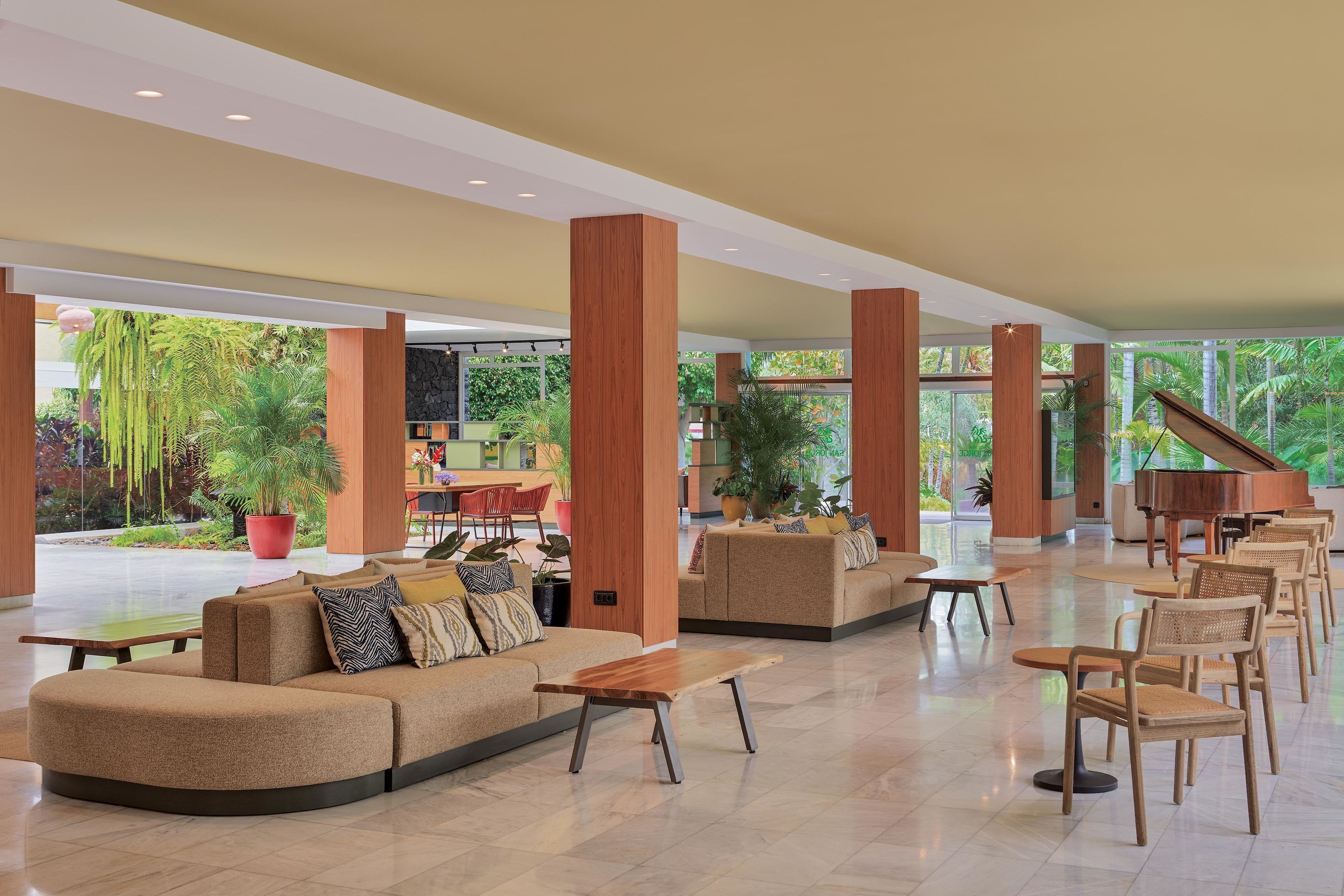 A spacious lobby with a curved sofa, wooden tables, and chairs, surrounded by large windows and potted plants.
