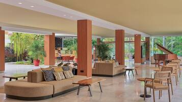 A spacious lobby with a curved sofa, wooden tables, and chairs, surrounded by large windows and potted plants.
