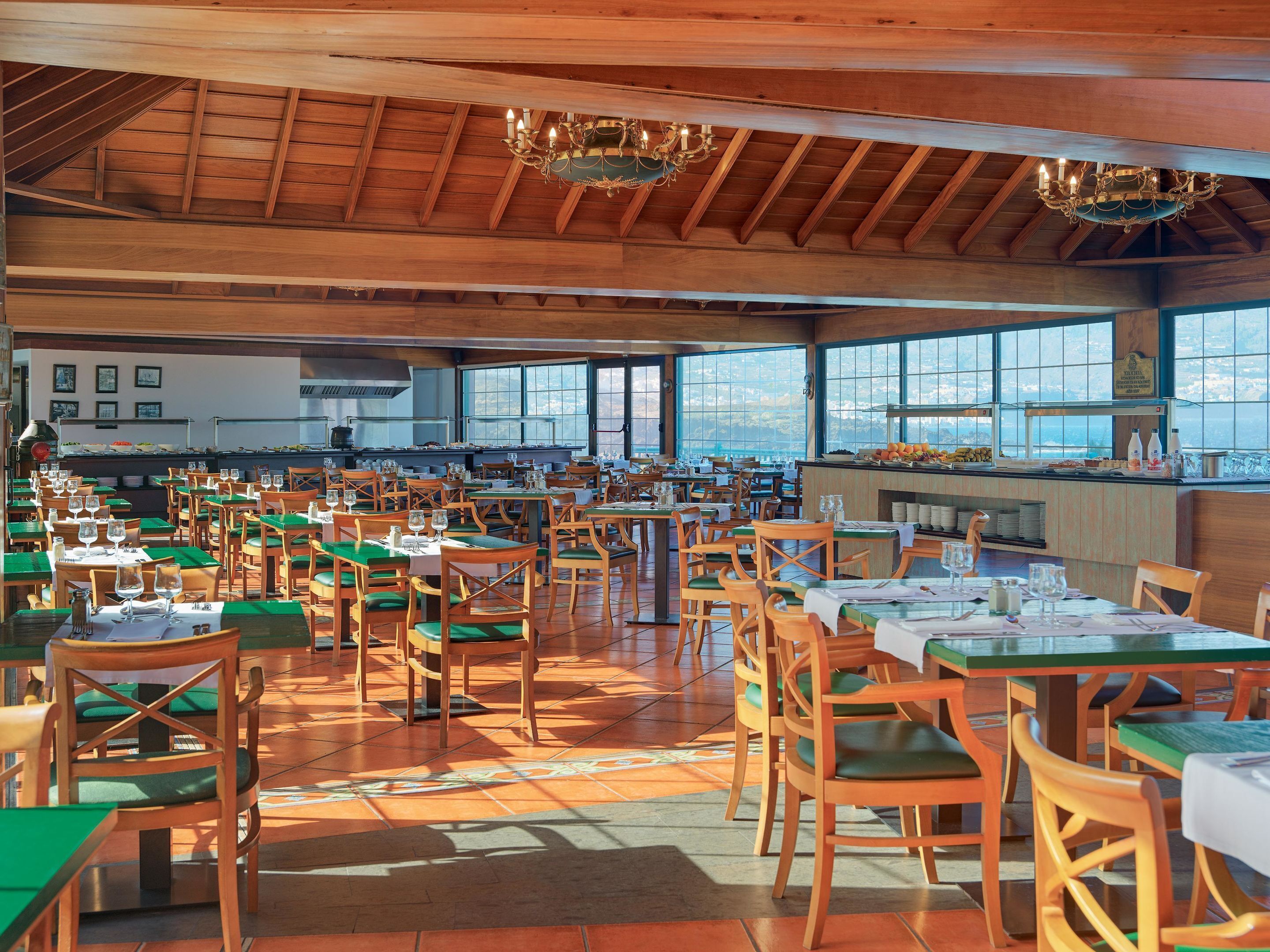 Un restaurant spacieux avec des tables et des chaises en bois, de grandes fenêtres et une cuisine visible au fond de la salle.