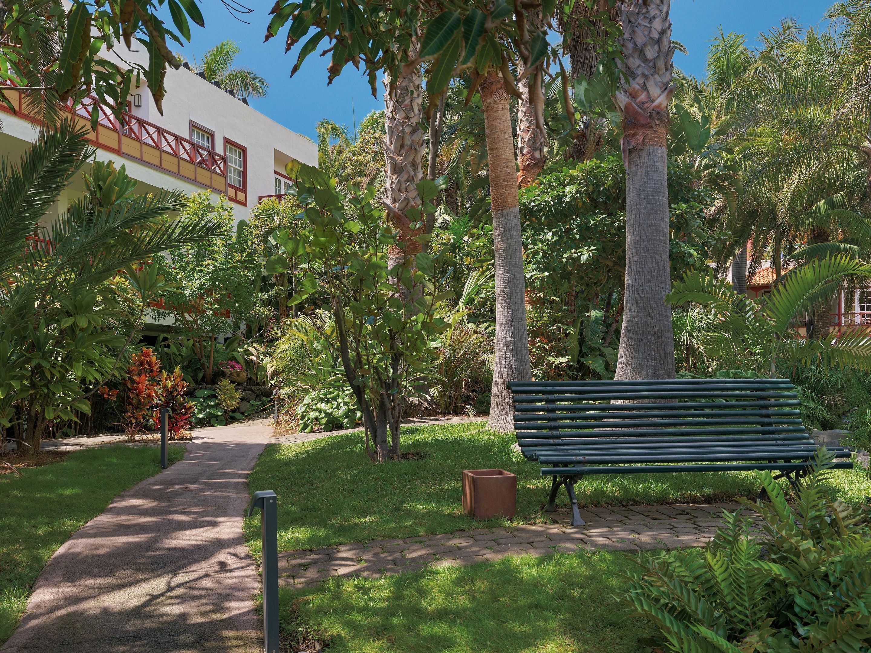 Un sendero conduce a un edificio de fachada blanca y detalles en rojo, rodeado de abundante vegetación y palmeras.