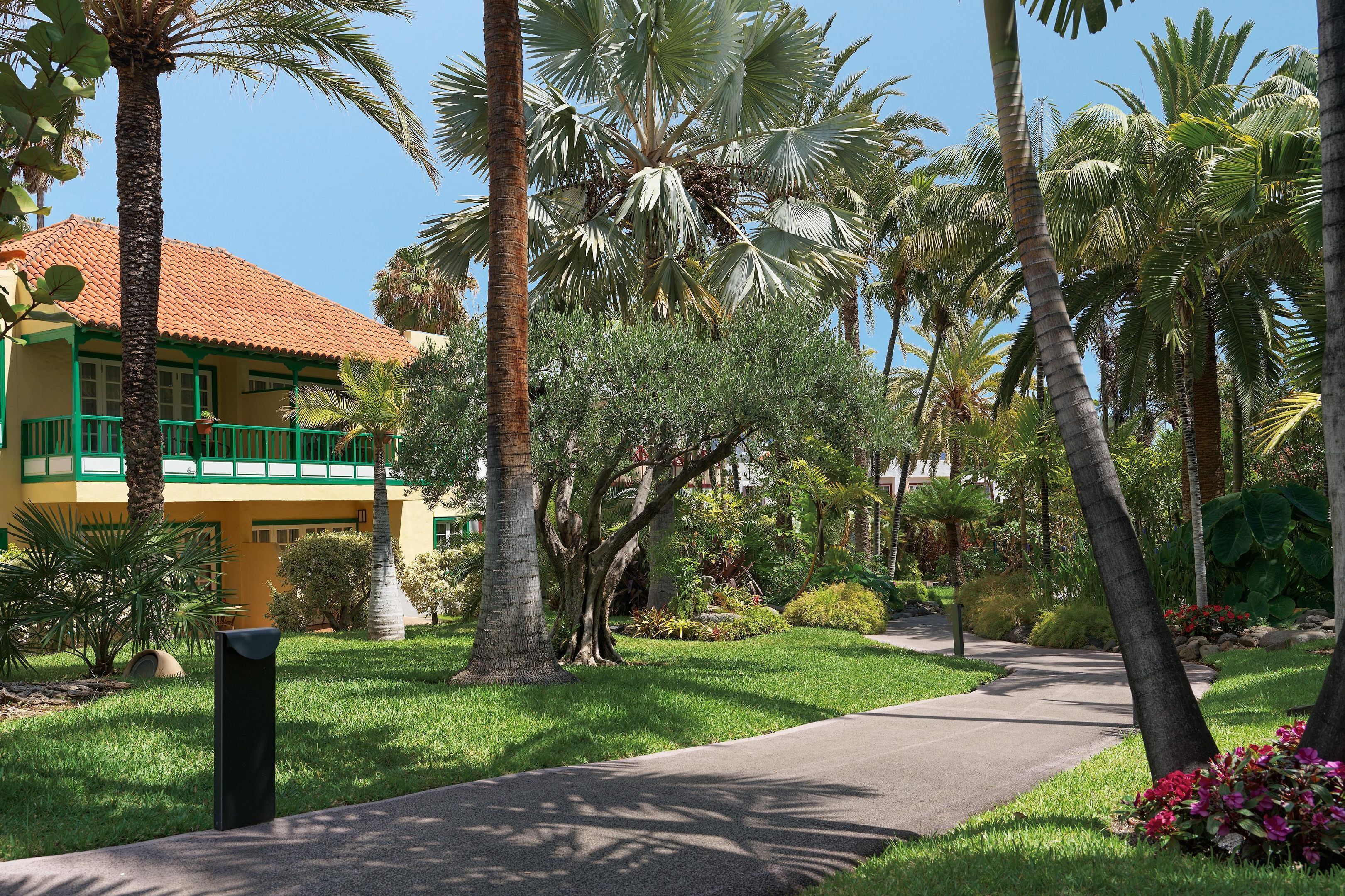 A pathway leading to a building with a red-tiled roof and green accents, surrounded by palm trees and lush greenery.