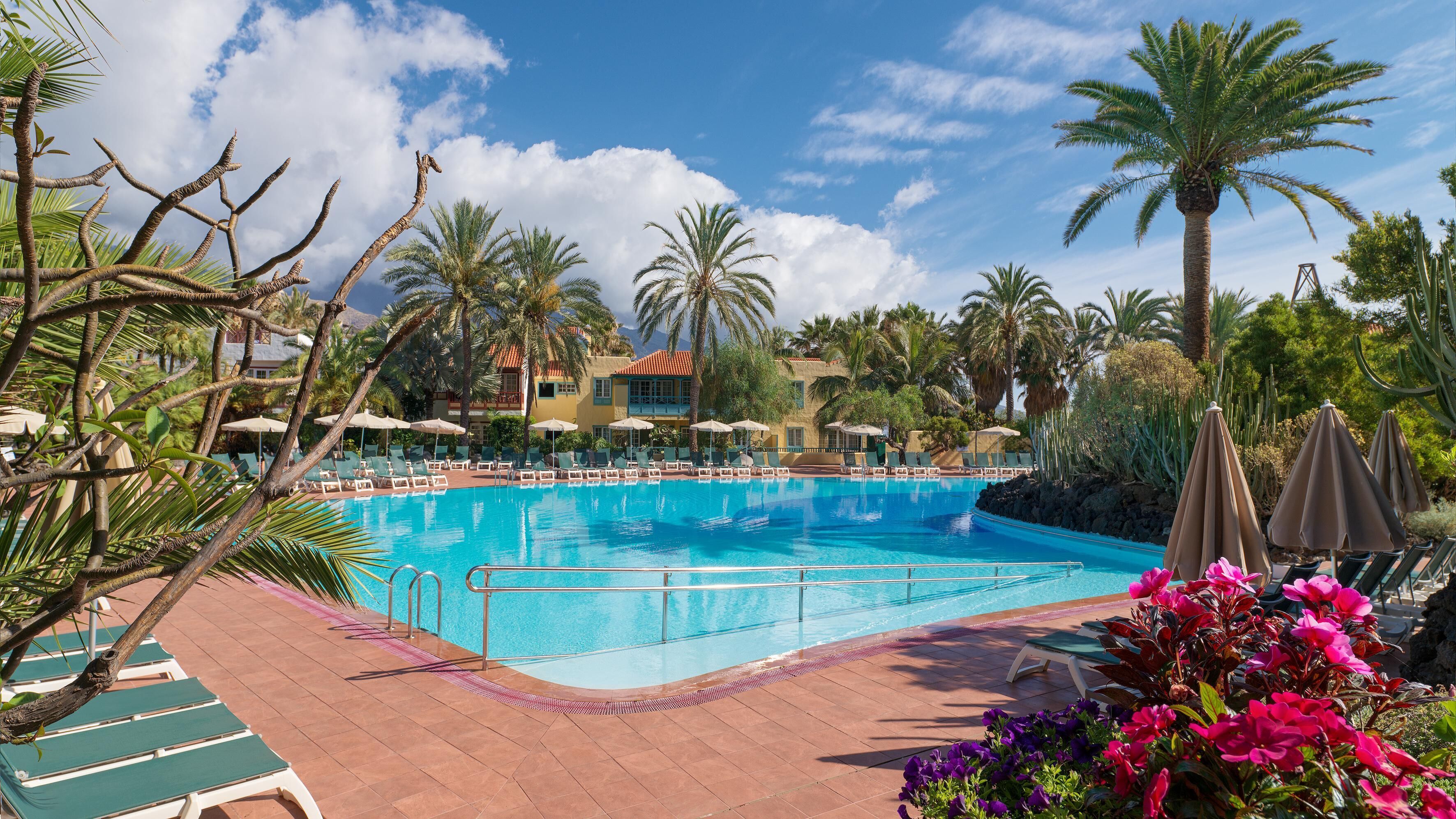 A pool area with lounge chairs and umbrellas, surrounded by palm trees and a building with a red roof.