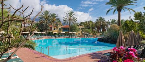 A pool area with lounge chairs and umbrellas, surrounded by palm trees and a building with a red roof.