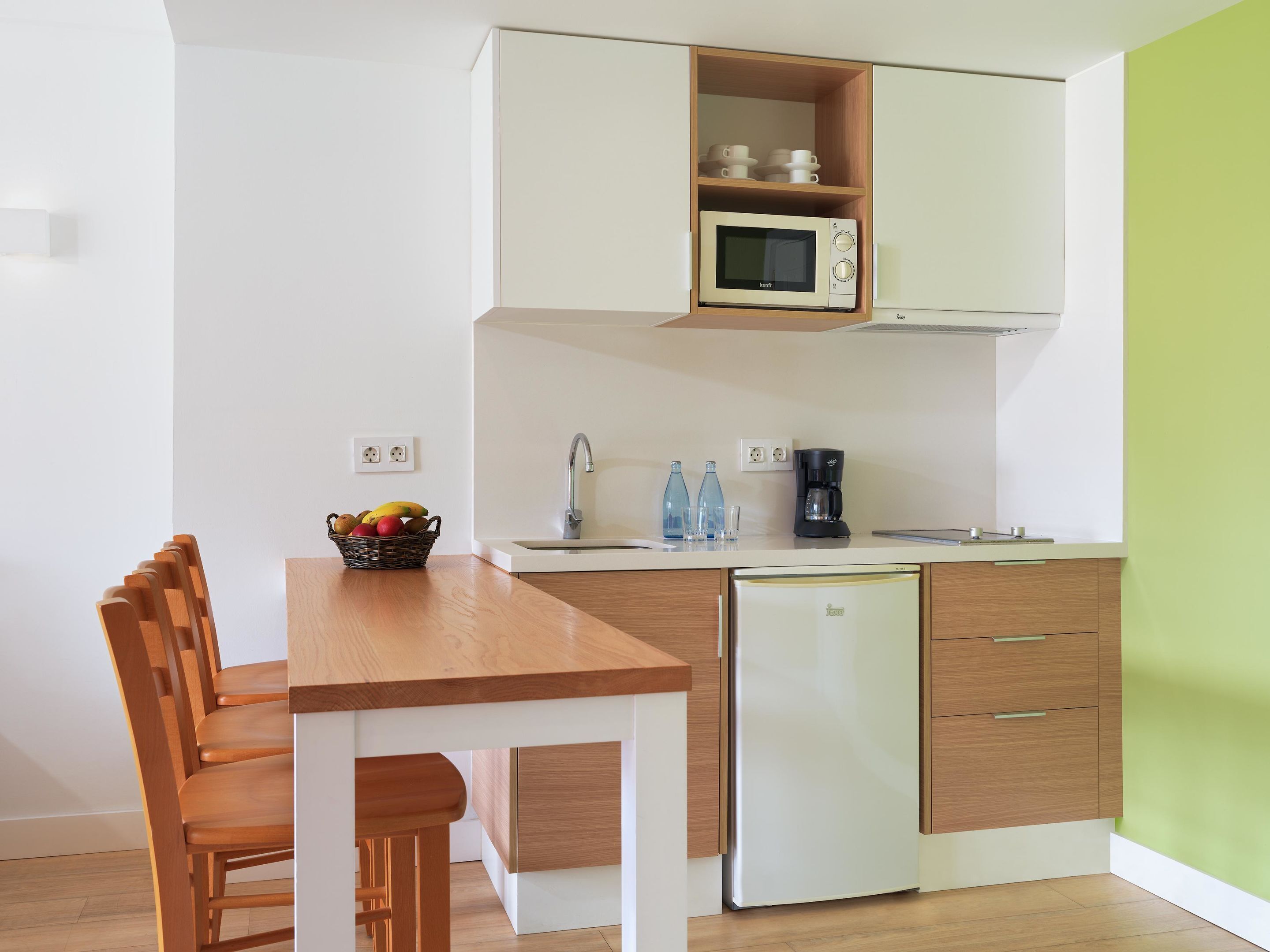 A compact kitchen with a microwave, coffee maker, and a small refrigerator. There is a wooden dining table with chairs and a fruit basket.