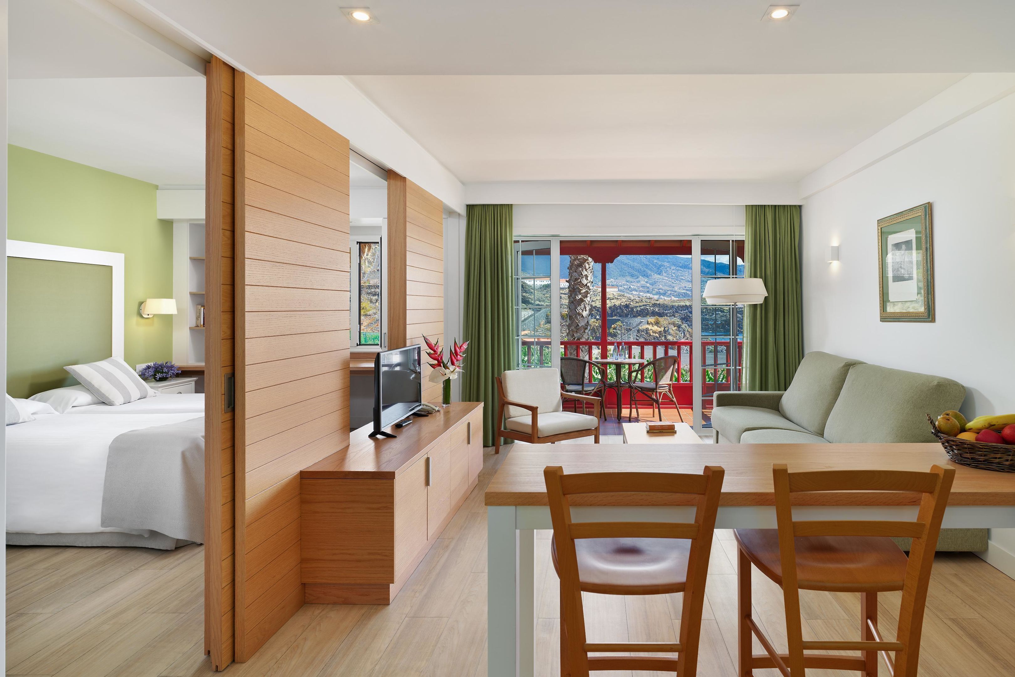 Quarto de hotel moderno com cama, escrivaninha com computador, área de refeições com cadeiras e varanda com vista para as montanhas.