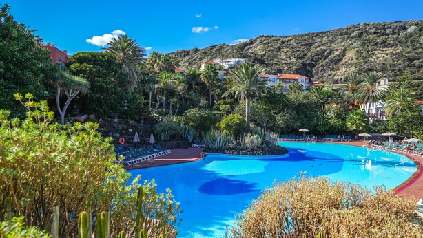 Una piscina rodeada de abundante vegetación, con montañas al fondo y edificios a lo lejos.