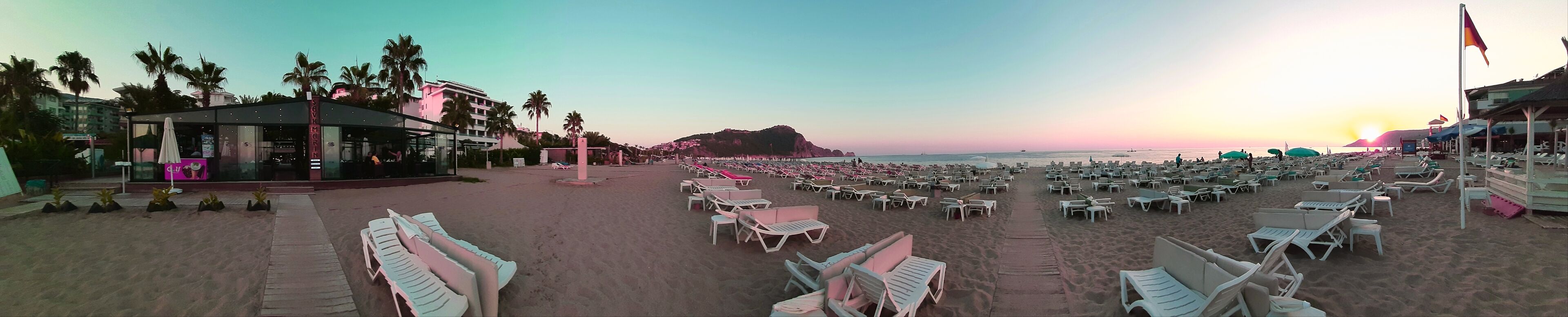 Una spiaggia con file di sedie a sdraio bianche e ombrelloni, un edificio con facciata in vetro e palme.