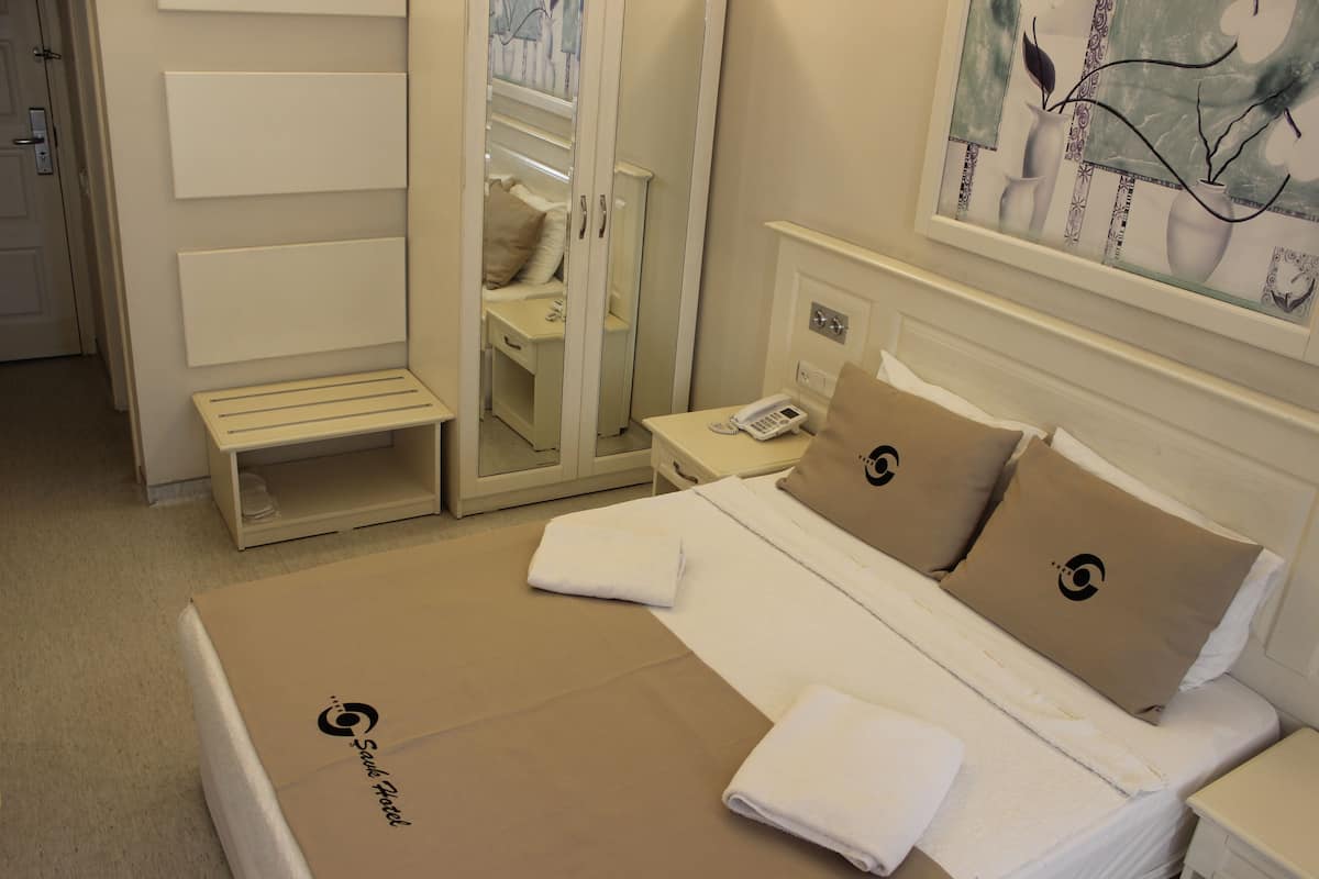 A hotel room with a bed, a bedside table, a nightstand, a mirror, a wardrobe, and a painting on the wall.