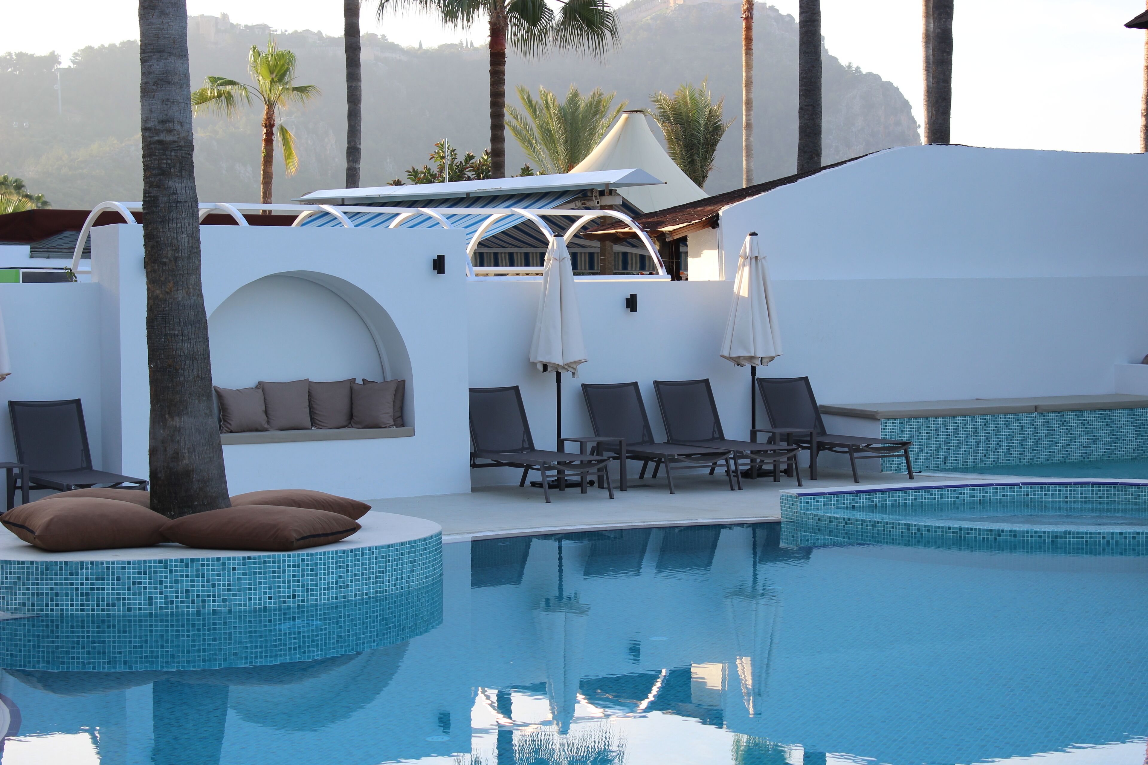 A pool area with lounge chairs and umbrellas, surrounded by palm trees and a white building with a tiled pool.