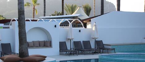A pool area with lounge chairs and umbrellas, surrounded by palm trees and a white building with a tiled pool.