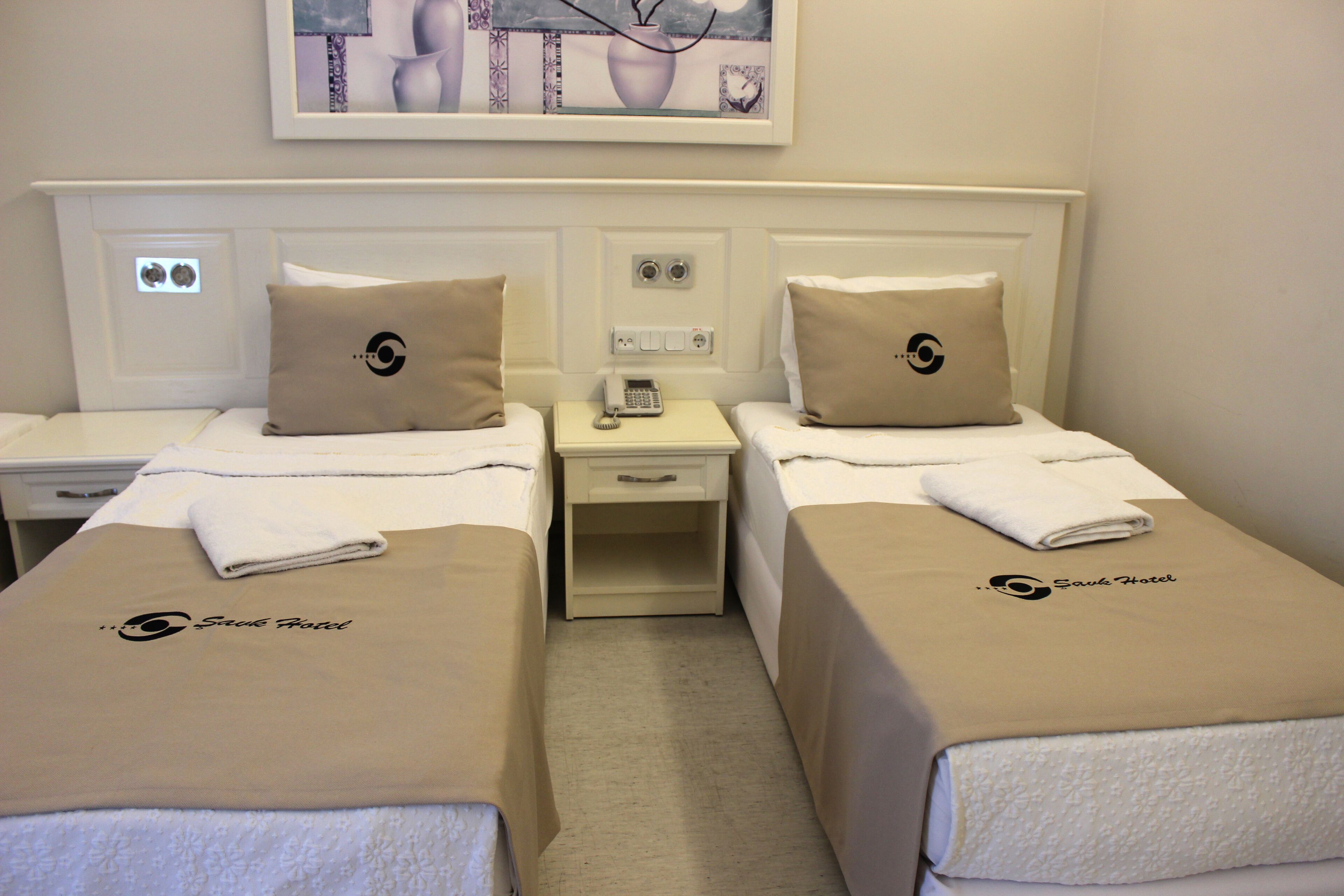 A hotel room with two beds, each with a beige bedspread and matching pillows, a small bedside table with a lamp, a wall-mounted painting, and a framed picture above the bed.
