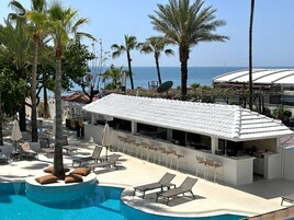 A resort with a pool, lounge chairs, and a bar area with a white tiled roof overlooking the sea.