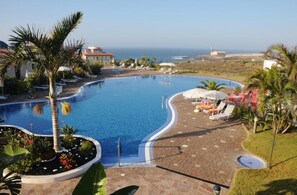 A pool with a curved edge, surrounded by lounge chairs and umbrellas, with a view of the sea and buildings in the background. - Luz del Mar (Los Silos)