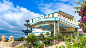 A hotel with a blue and white facade, a fountain, and lush greenery in the foreground. - Daima Biz Hotel - Dolusu Aquapark Access - All inclusive (Kemer)