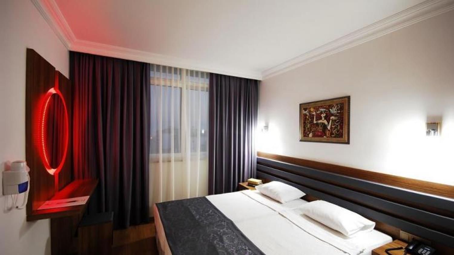Ein Hotelzimmer mit Bett, Nachttisch, Wandlampe, einem Bild und einem Fenster mit Vorhängen.