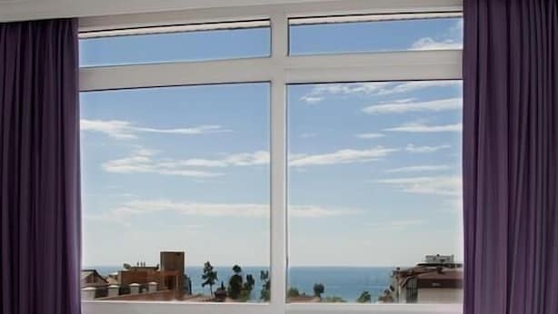 A bedroom with a large window, a bed, and a view of the sea and buildings. - Sealine Hotel (Alanya)
