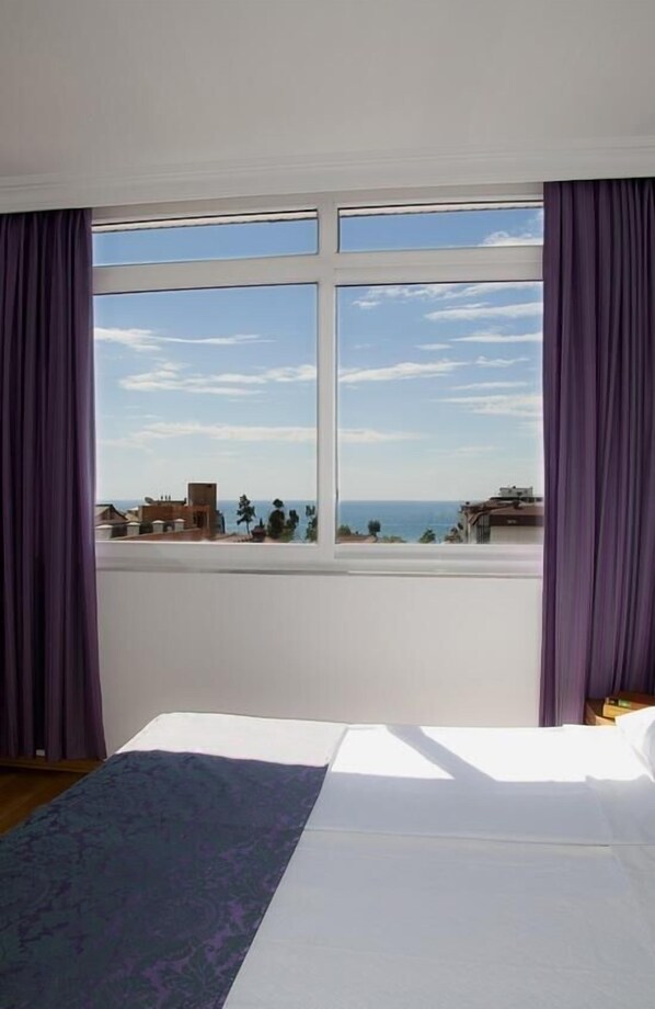A bedroom with a large window, a bed, and a view of the sea and buildings. - Sealine Hotel (Alanya)