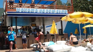 A beach club with a sign reading "HAVANA BEACH CLUB". There are people lounging on sunbeds and chairs, and a lifeguard tower is visible. - Sealine Hotel (Alanya)