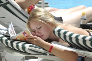 A person lying on a striped lounge chair by a pool, reading a magazine. - Sealine Hotel (Alanya)